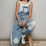 Denim coveralls with patches and a white tank top on model with hat on stool