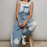 Denim coveralls with patches and a white tank top on model with hat on stool