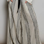 A person wearing loose, wide-leg pleated pants in a light beige and gray striped textured fabric, styled with a black ribbon tie belt at the waist, a white long-sleeve top, and black shoes.