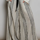 A person wearing loose, wide-leg pleated pants in a light beige and gray striped textured fabric, styled with a black ribbon tie belt at the waist, a white long-sleeve top, and black shoes.