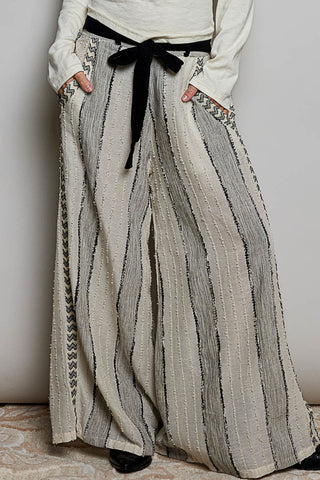 A person wearing loose, wide-leg pleated pants in a light beige and gray striped textured fabric, styled with a black ribbon tie belt at the waist, a white long-sleeve top, and black shoes.
