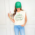 Woman wearing a white sleeveless shirt with text, blue jeans, and a green cap, holding a drink.