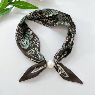 A square silk scarf with a dark brown base and a green, beige, and white botanical pattern, tied in a knot at the center with a white faux-pearl accent, displayed on a light gray surface with green leaves in the top-left corner.