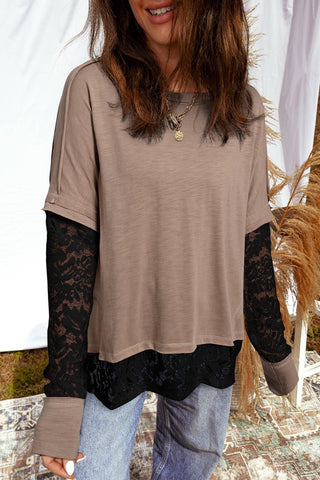 Person wearing a beige long-sleeve top with lace sleeves and jeans, standing against a white curtain.