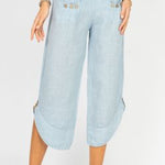 A woman standing, wearing white linen pants with side ties and a white top, paired with a blue denim jacket and blue strappy sandals.