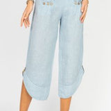 A woman standing, wearing white linen pants with side ties and a white top, paired with a blue denim jacket and blue strappy sandals.