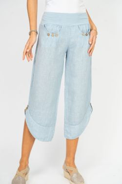 A woman standing, wearing white linen pants with side ties and a white top, paired with a blue denim jacket and blue strappy sandals.