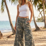 Woman in white tank top and camouflage pants standing on a beach with palm trees.