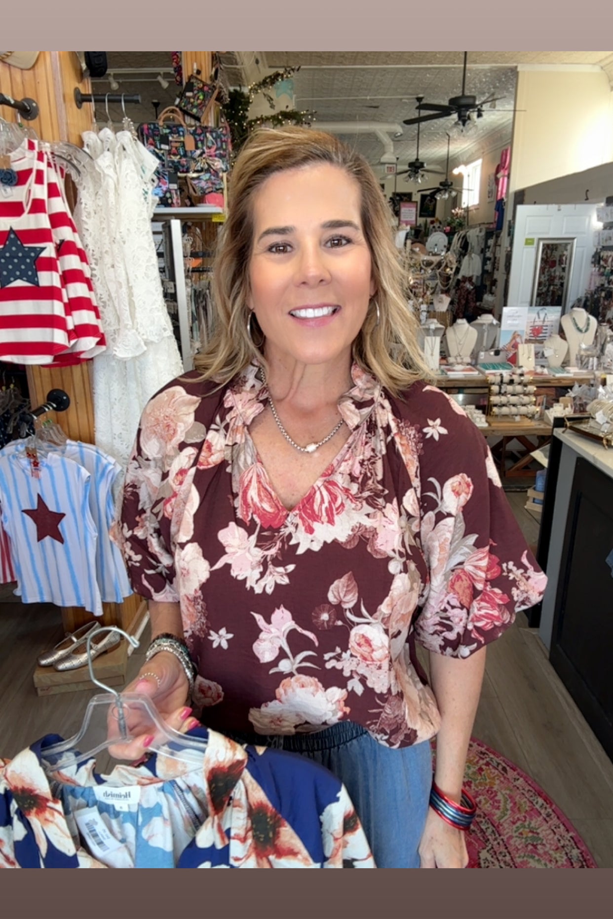 Woman wearing a floral blouse in a store setting with clothing racks and an American flag in the background.