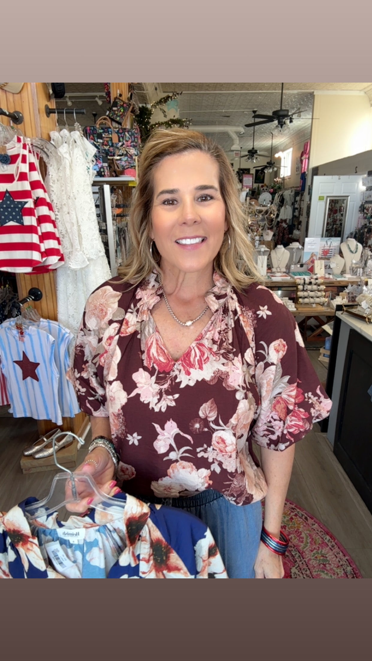 Woman wearing a floral blouse in a store setting with clothing racks and an American flag in the background.