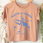 Beige t-shirt with blue graphic and text 'Meet Me at the Beach' on a white wall with decorative elements.