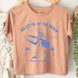 Beige t-shirt with blue graphic and text 'Meet Me at the Beach' on a white wall with decorative elements.