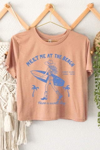 Beige t-shirt with blue graphic and text 'Meet Me at the Beach' on a white wall with decorative elements.