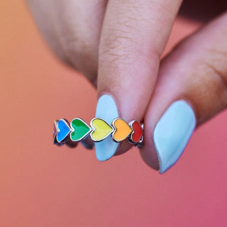 Ring with rainbow heart-shaped stones on a hand with light blue nail polish against a blurred background