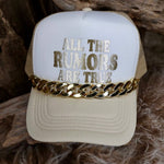 Beige cap with 'All the rumors are true' text and gold chain on a wooden surface