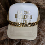 Beige cap with 'All the rumors are true' text and gold chain on a wooden surface