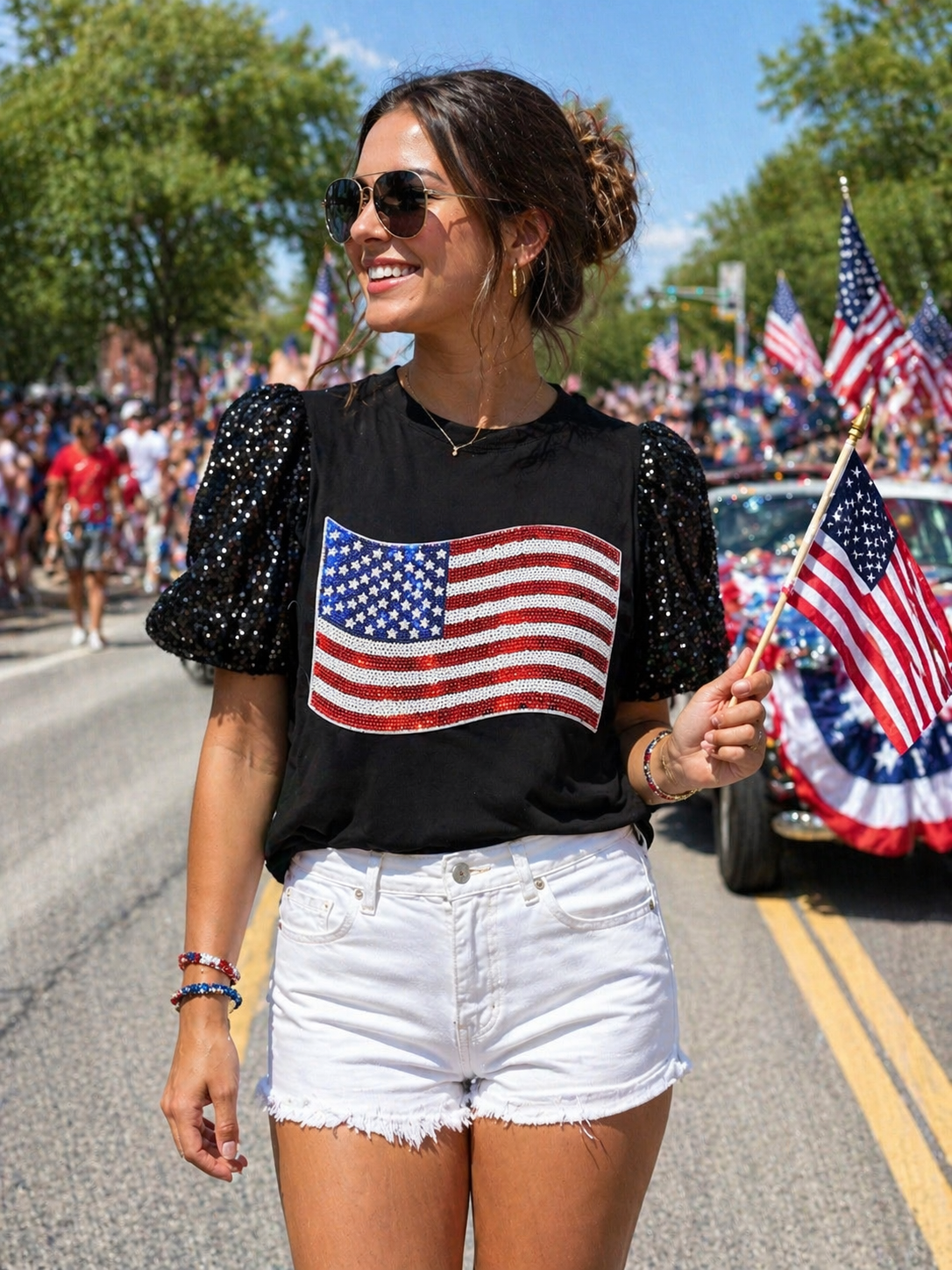 Woman wearing a black t-shirt with an American flag design, white shorts, and sunglasses, standing on a street with American flags in the background.