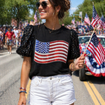 Woman wearing a black t-shirt with an American flag design, white shorts, and sunglasses, standing on a street with American flags in the background.