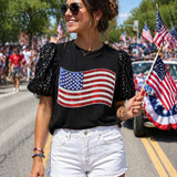 Woman wearing a black t-shirt with an American flag design, white shorts, and sunglasses, standing on a street with American flags in the background.