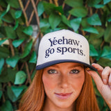 Person wearing a cap with 'Yeehaw, go sports.' text against a green leafy background
