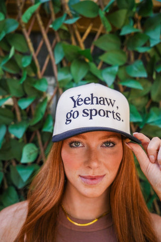 Person wearing a cap with 'Yeehaw, go sports.' text against a green leafy background