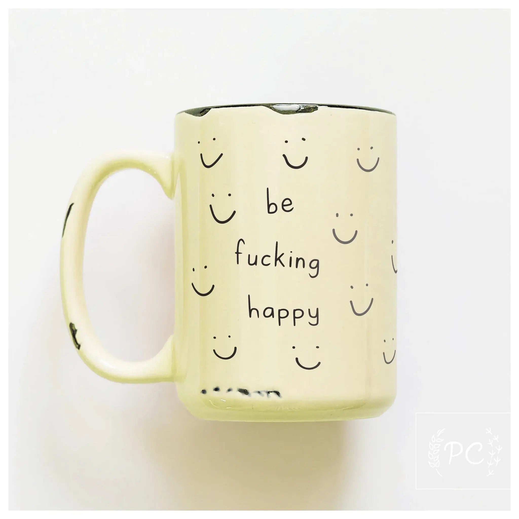 Yellow mug with smiley faces and text 'be fucking happy' on a white background