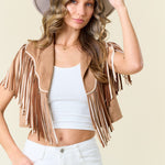 Woman wearing a beige fringe jacket, white top, and white pants against a beige background