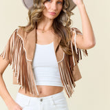 Woman wearing a beige fringe jacket, white top, and white pants against a beige background