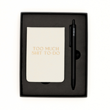 Notebook with 'Too Much Shit To Do' text and a pen in a black box on a white background
