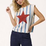 Woman wearing a sleeveless top with a red star and blue and white striped pattern, paired with dark pants on a white background.