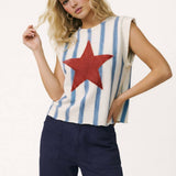 Woman wearing a sleeveless top with a red star and blue and white striped pattern, paired with dark pants on a white background.