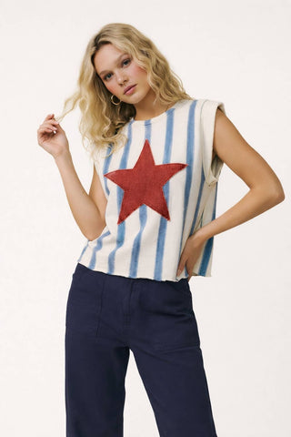 Woman wearing a sleeveless top with a red star and blue and white striped pattern, paired with dark pants on a white background.