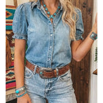 Woman wearing a denim shirt and jeans with a belt, standing in a room with wooden walls.