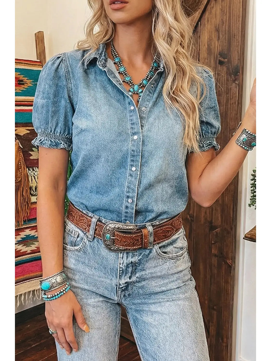 Woman wearing a denim shirt and jeans with a belt, standing in a room with wooden walls.