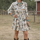 Woman in a patterned dress and cowboy boots standing in an outdoor setting with trees and a red building.
