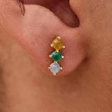 Close-up of an ear with a gold earring featuring three colored gemstones.