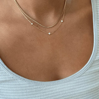 Close-up of a person wearing a gold necklace with small round pendants on a light gray top.