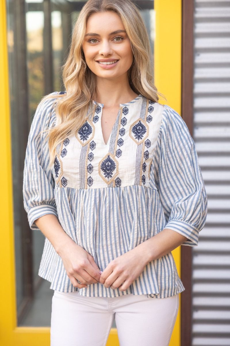 Woman wearing a blue and white striped blouse with embroidery in front of a yellow and gray background.