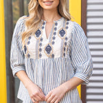 Woman wearing a blue and white striped blouse with embroidery in front of a yellow and gray background.