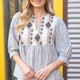 Woman wearing a blue and white striped blouse with embroidery in front of a yellow and gray background.