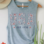 Blue tank top with 'USA EST. 1776' design on a hanger against a white wall.