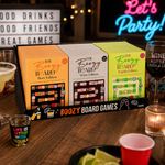 A boxed set of three 'The Boozy Board Games' displayed on a wooden bar table with drinks, cups, and party decor in the background. The set includes Beer Edition, Wine Edition, and Expi Edition boxes.