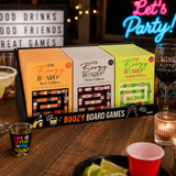 A boxed set of three 'The Boozy Board Games' displayed on a wooden bar table with drinks, cups, and party decor in the background. The set includes Beer Edition, Wine Edition, and Expi Edition boxes.