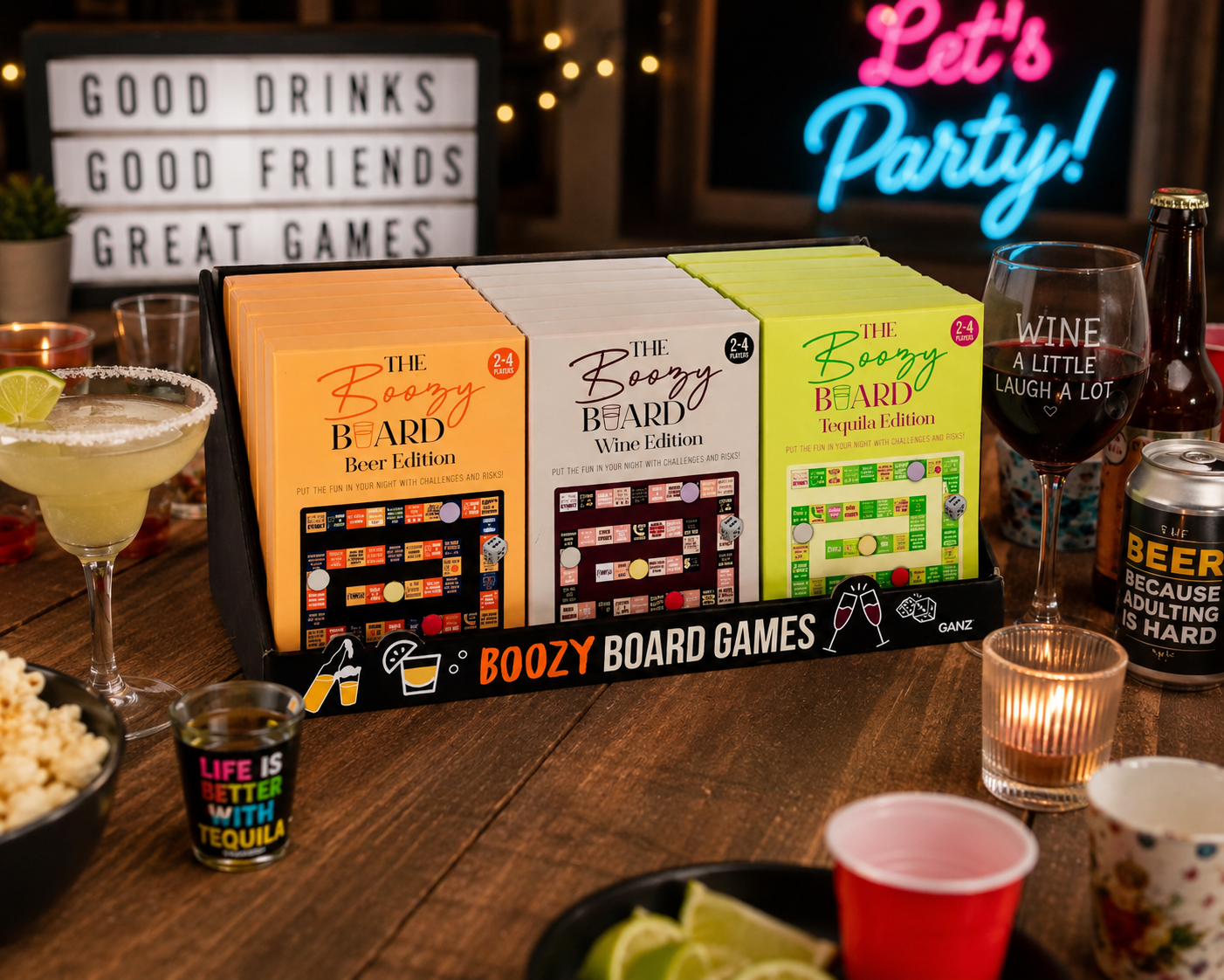 A boxed set of three 'The Boozy Board Games' displayed on a wooden bar table with drinks, cups, and party decor in the background. The set includes Beer Edition, Wine Edition, and Expi Edition boxes.