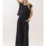 A woman modeling a black sleeveless jumpsuit with a loose top and wide-leg pants, paired with beige flat sandals, against a plain light background.