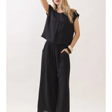 A woman modeling a black sleeveless jumpsuit with a loose top and wide-leg pants, paired with beige flat sandals, against a plain light background.