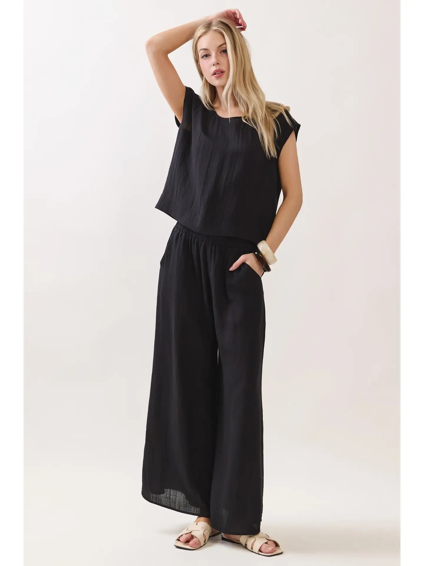 A woman modeling a black sleeveless jumpsuit with a loose top and wide-leg pants, paired with beige flat sandals, against a plain light background.