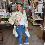 Woman standing in a store holding a decorative item, wearing a floral jacket and blue jeans.