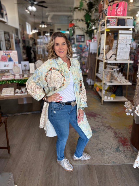 Woman standing in a store holding a decorative item, wearing a floral jacket and blue jeans.