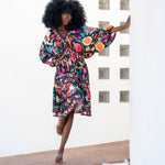 Woman in a colorful dress standing on a wooden deck with a white wall and geometric patterned wall in the background.
